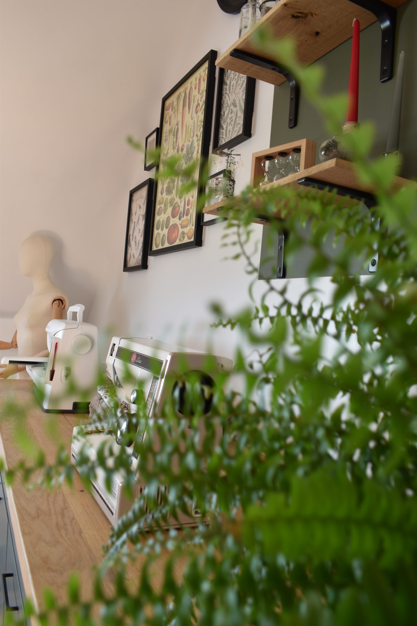 posé sur un plan de travail : un mannequin de couture (bras en bois), une brodeuse et une machine à coudre au travers d'une fougère